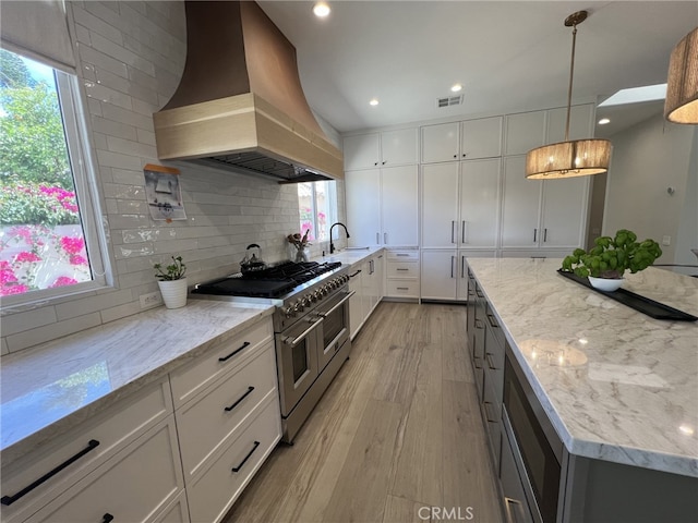 kitchen featuring decorative light fixtures, light stone countertops, range with two ovens, custom range hood, and white cabinets