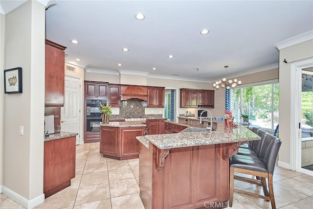 kitchen with a kitchen bar, custom exhaust hood, black double oven, decorative light fixtures, and a center island with sink