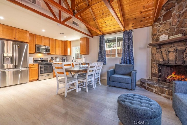 kitchen with a stone fireplace, vaulted ceiling with beams, appliances with stainless steel finishes, light hardwood / wood-style floors, and wood ceiling