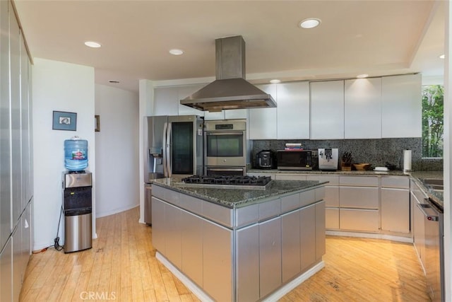 kitchen with a center island, light wood-type flooring, appliances with stainless steel finishes, island exhaust hood, and decorative backsplash