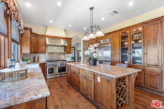 kitchen featuring premium appliances, tasteful backsplash, an island with sink, hanging light fixtures, and light stone counters