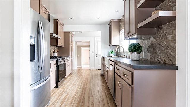 kitchen with wall chimney range hood, sink, appliances with stainless steel finishes, tasteful backsplash, and light hardwood / wood-style floors