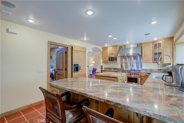 kitchen featuring wall chimney range hood, sink, appliances with stainless steel finishes, a kitchen island, and light stone counters