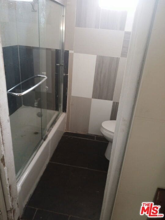 bathroom featuring tile walls, tile floors, bath / shower combo with glass door, and toilet