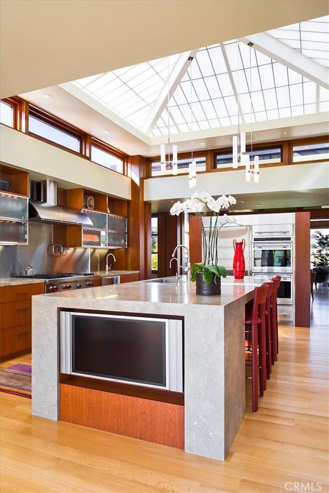 kitchen featuring sink, wall chimney range hood, a large island with sink, decorative light fixtures, and light wood-type flooring