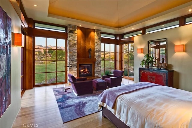 bedroom featuring light wood-type flooring, a raised ceiling, multiple windows, and a stone fireplace