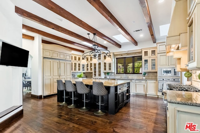 kitchen featuring appliances with stainless steel finishes, beamed ceiling, an island with sink, and cream cabinets