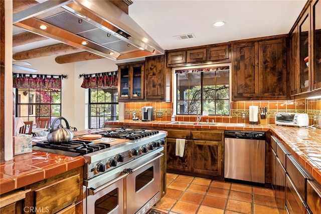 kitchen with wall chimney range hood, stainless steel appliances, tile countertops, backsplash, and sink