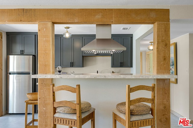 kitchen with gray cabinetry, wall chimney exhaust hood, a kitchen bar, and stainless steel refrigerator