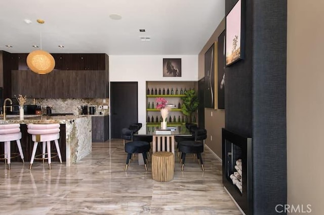 kitchen with dark brown cabinetry, decorative light fixtures, decorative backsplash, light stone counters, and a breakfast bar area