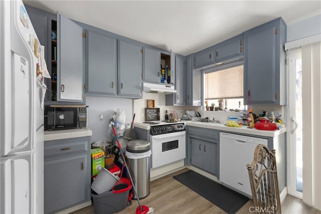 kitchen with under cabinet range hood, white appliances, gray cabinets, backsplash, and light wood finished floors