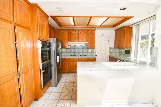 kitchen featuring backsplash, stainless steel appliances, a raised ceiling, kitchen peninsula, and tile counters