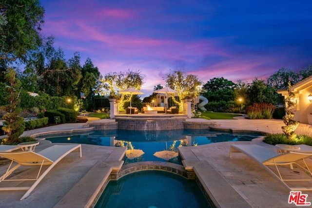 pool at dusk with a patio area and an in ground hot tub