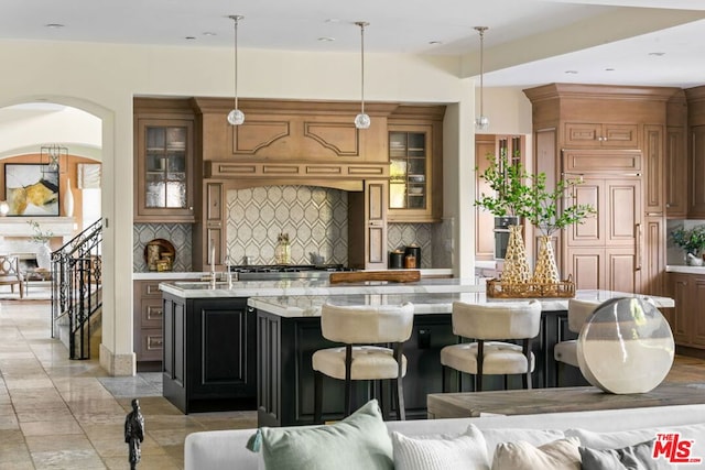 kitchen with decorative backsplash, hanging light fixtures, an island with sink, light stone counters, and a breakfast bar area