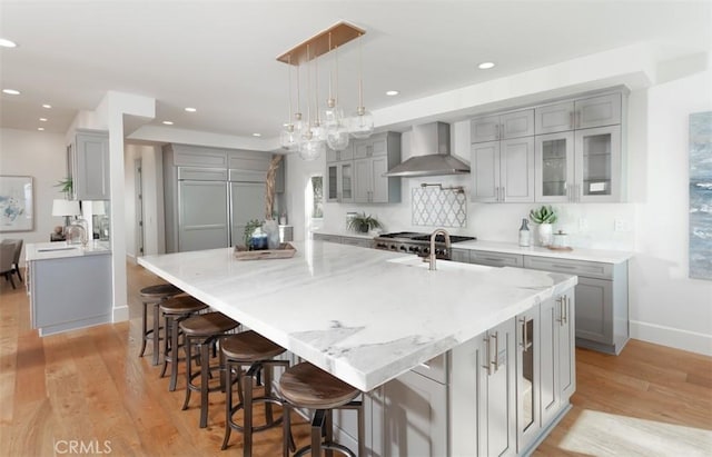 kitchen with light wood-type flooring, wall chimney exhaust hood, a large island with sink, gray cabinets, and hanging light fixtures