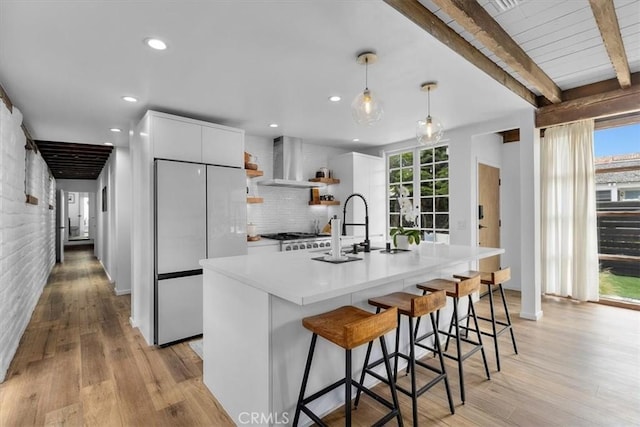 kitchen featuring built in fridge, white cabinets, decorative light fixtures, wall chimney range hood, and beam ceiling