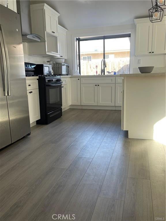 kitchen with stainless steel refrigerator, wall chimney exhaust hood, hanging light fixtures, black gas range oven, and white cabinets