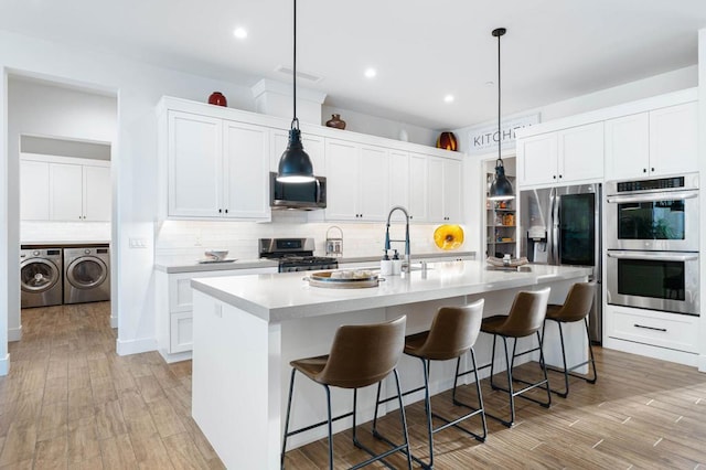 kitchen featuring tasteful backsplash, an island with sink, washer and dryer, white cabinetry, and appliances with stainless steel finishes