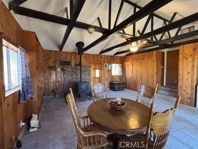 carpeted dining room with wooden walls, lofted ceiling with beams, and a wood stove