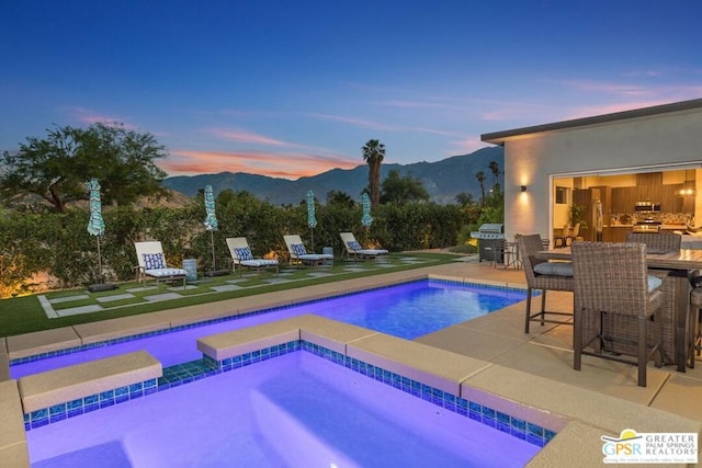 pool at dusk with a mountain view, area for grilling, a patio, and an in ground hot tub