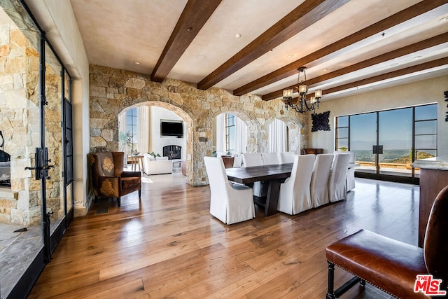 dining space featuring a stone fireplace, beam ceiling, hardwood / wood-style floors, and a chandelier