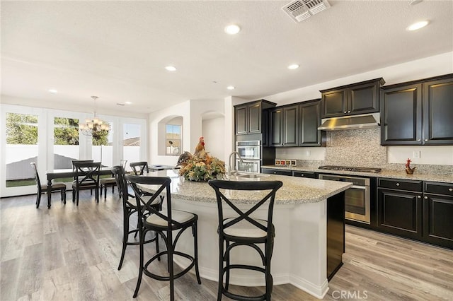 kitchen featuring an island with sink, light wood-type flooring, a kitchen breakfast bar, hanging light fixtures, and light stone countertops