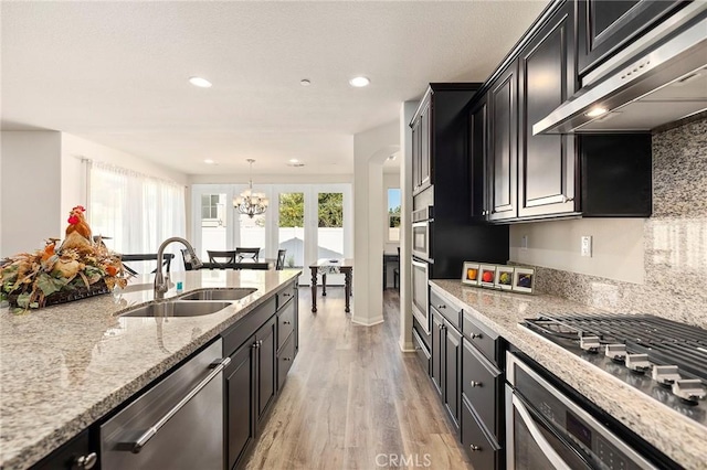 kitchen with range hood, sink, an inviting chandelier, light stone countertops, and stainless steel appliances