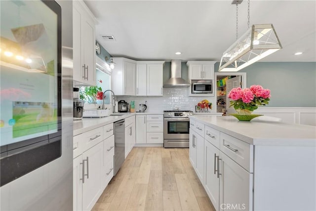 kitchen with white cabinets, wall chimney range hood, appliances with stainless steel finishes, decorative light fixtures, and light hardwood / wood-style floors