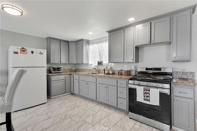 kitchen featuring gray cabinetry, sink, stainless steel range with gas cooktop, light stone counters, and white refrigerator