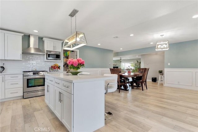 kitchen with white cabinets, wall chimney range hood, hanging light fixtures, appliances with stainless steel finishes, and light hardwood / wood-style floors