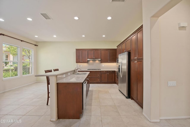 kitchen featuring light tile flooring, stainless steel appliances, a kitchen breakfast bar, a center island with sink, and light stone countertops