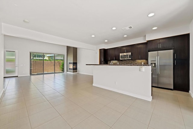 kitchen with stainless steel appliances, decorative backsplash, a kitchen island with sink, light tile patterned flooring, and dark brown cabinetry