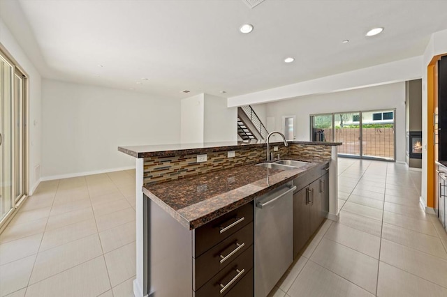 kitchen with stainless steel dishwasher, light tile patterned floors, a kitchen island with sink, dark stone countertops, and sink