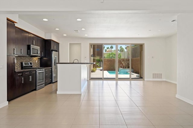 kitchen featuring a center island with sink, stainless steel appliances, decorative backsplash, dark brown cabinetry, and light tile patterned flooring