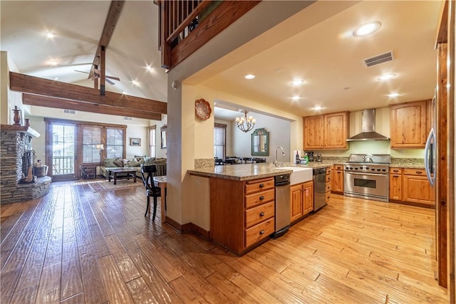 kitchen featuring wall chimney range hood, light hardwood / wood-style flooring, appliances with stainless steel finishes, beam ceiling, and kitchen peninsula