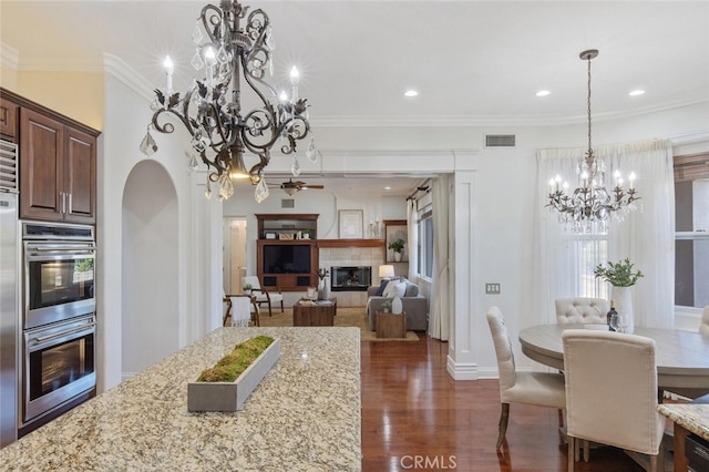 kitchen with ornamental molding, dark hardwood / wood-style flooring, light stone countertops, stainless steel double oven, and a tiled fireplace