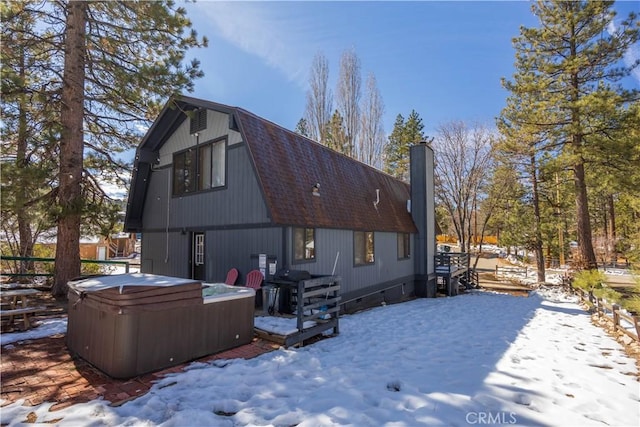 snow covered rear of property with a hot tub, a chimney, a shingled roof, and a gambrel roof