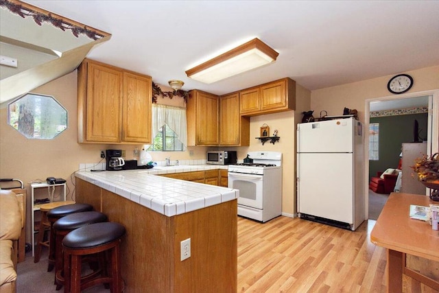 kitchen with white appliances, kitchen peninsula, light hardwood / wood-style flooring, tile counters, and a kitchen breakfast bar