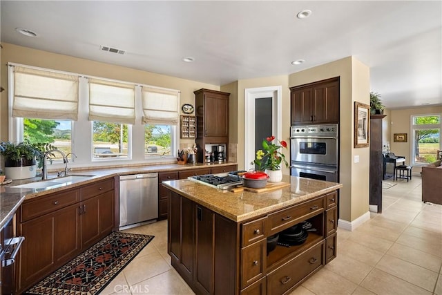 kitchen with dark brown cabinetry, light tile patterned floors, light stone counters, a kitchen island, and appliances with stainless steel finishes