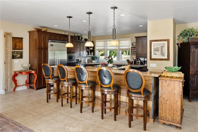 kitchen with hanging light fixtures, built in appliances, light stone countertops, dark brown cabinets, and a breakfast bar area