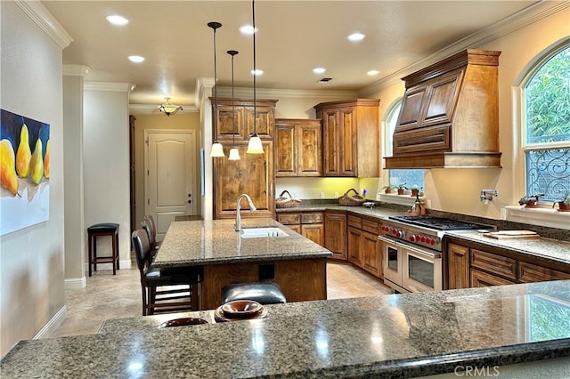 kitchen featuring an island with sink, double oven range, dark stone countertops, custom range hood, and sink