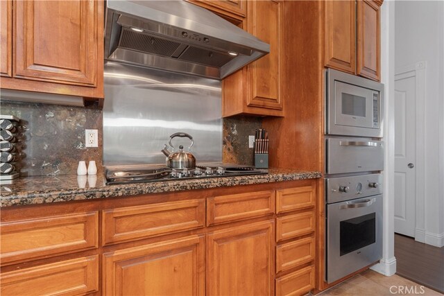 kitchen featuring wall chimney range hood, light hardwood / wood-style flooring, stainless steel appliances, dark stone counters, and backsplash