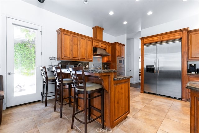 kitchen featuring dark stone counters, built in appliances, a breakfast bar area, and light tile floors