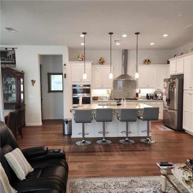 kitchen featuring a breakfast bar, stainless steel appliances, a kitchen island with sink, wall chimney range hood, and pendant lighting