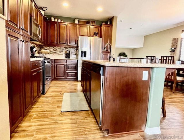 kitchen featuring a kitchen island with sink, stainless steel appliances, a kitchen bar, light wood-type flooring, and tasteful backsplash