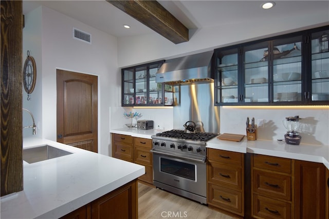 kitchen with beam ceiling, stainless steel range, wall chimney exhaust hood, sink, and light hardwood / wood-style floors