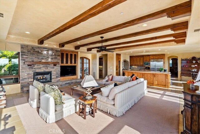 living room featuring ceiling fan, beam ceiling, and a stone fireplace