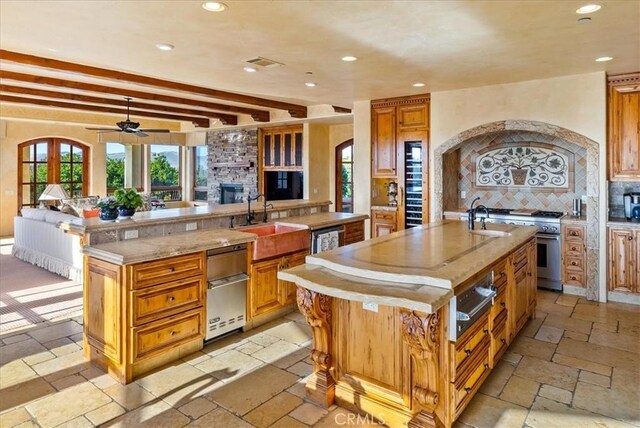 kitchen featuring sink, tasteful backsplash, a stone fireplace, beamed ceiling, and an island with sink