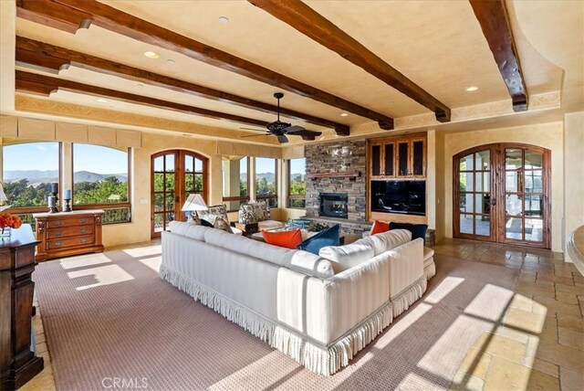 living room featuring a mountain view, french doors, a stone fireplace, ceiling fan, and beamed ceiling