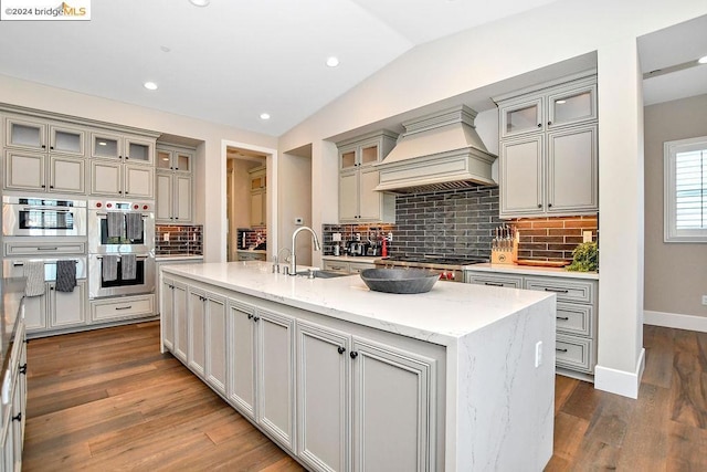 kitchen featuring backsplash, sink, an island with sink, light stone counters, and custom range hood